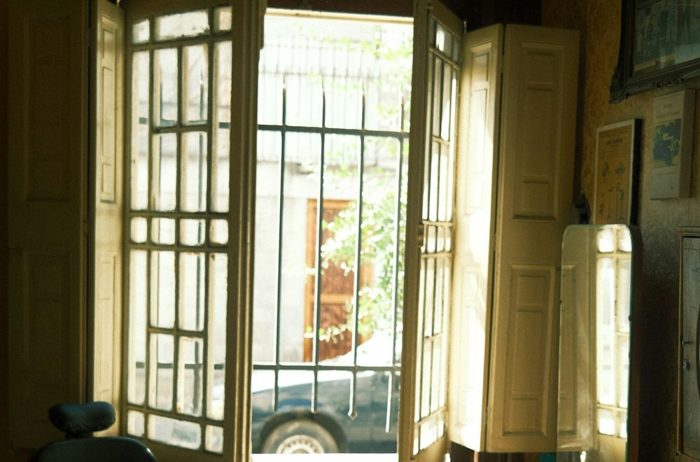 The doorway of the shop standing open onto the street at midday, with a barber chair inside by the window